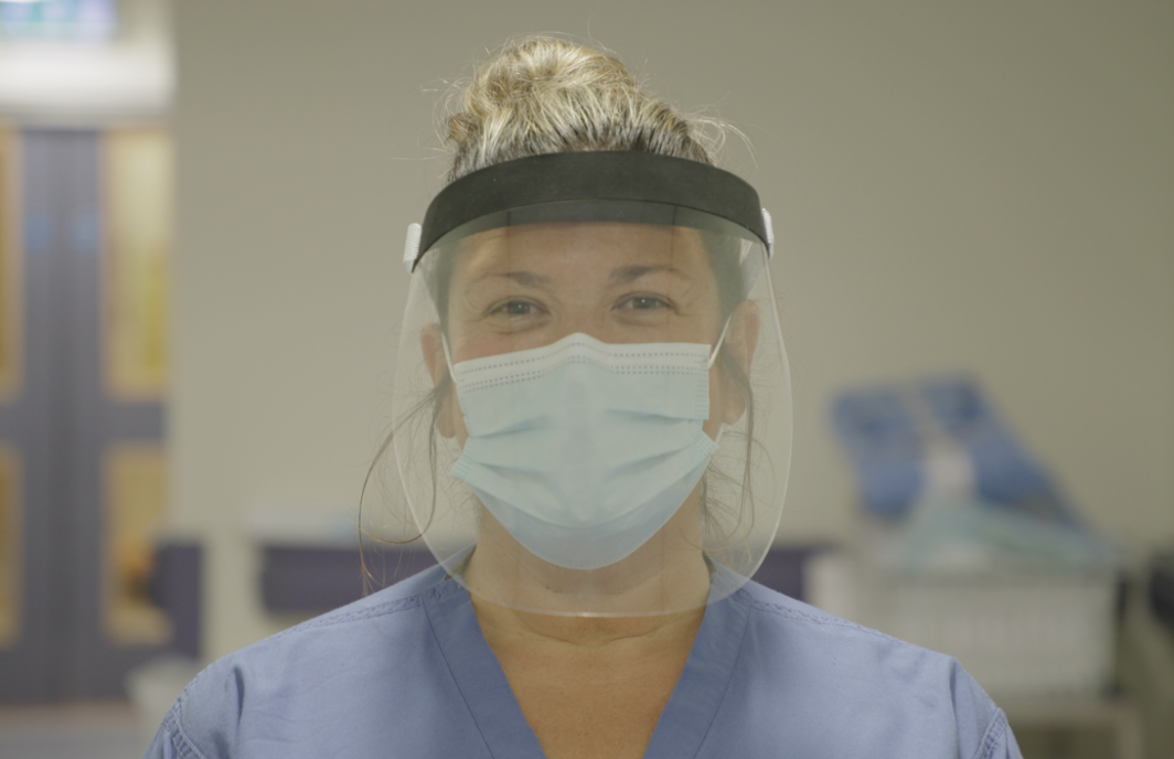 A clinical member of staff wearing blue scrubs, a surgical mask and a face shield smiles at the camera
