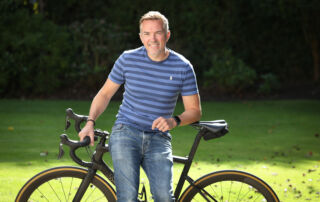 A white man with short hair and a blue striped T-shirt and jeans leans against a black bike with a green lawn behind him