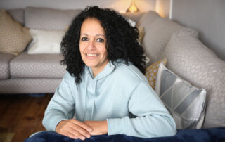 A lady with black curly hair in a pale blue hooded top sits on a silver corner sofa with cushions