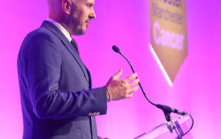 A man with a shaven head and smart suit stands on stage at a lectern