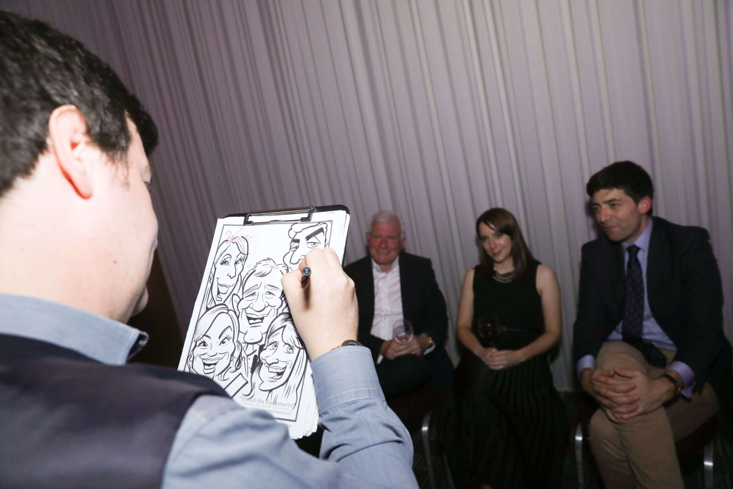 The back of a man's head he holds a drawing pad showing a partly finished caricature two men and a woman (centre) sit smiling in front of him