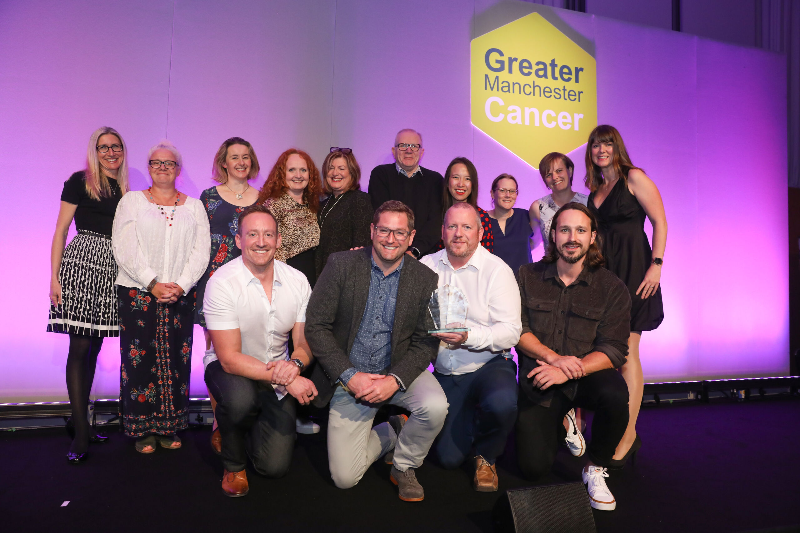 A group of men and women stand and crouch on stage as they celebrate at GM Cancer Awards