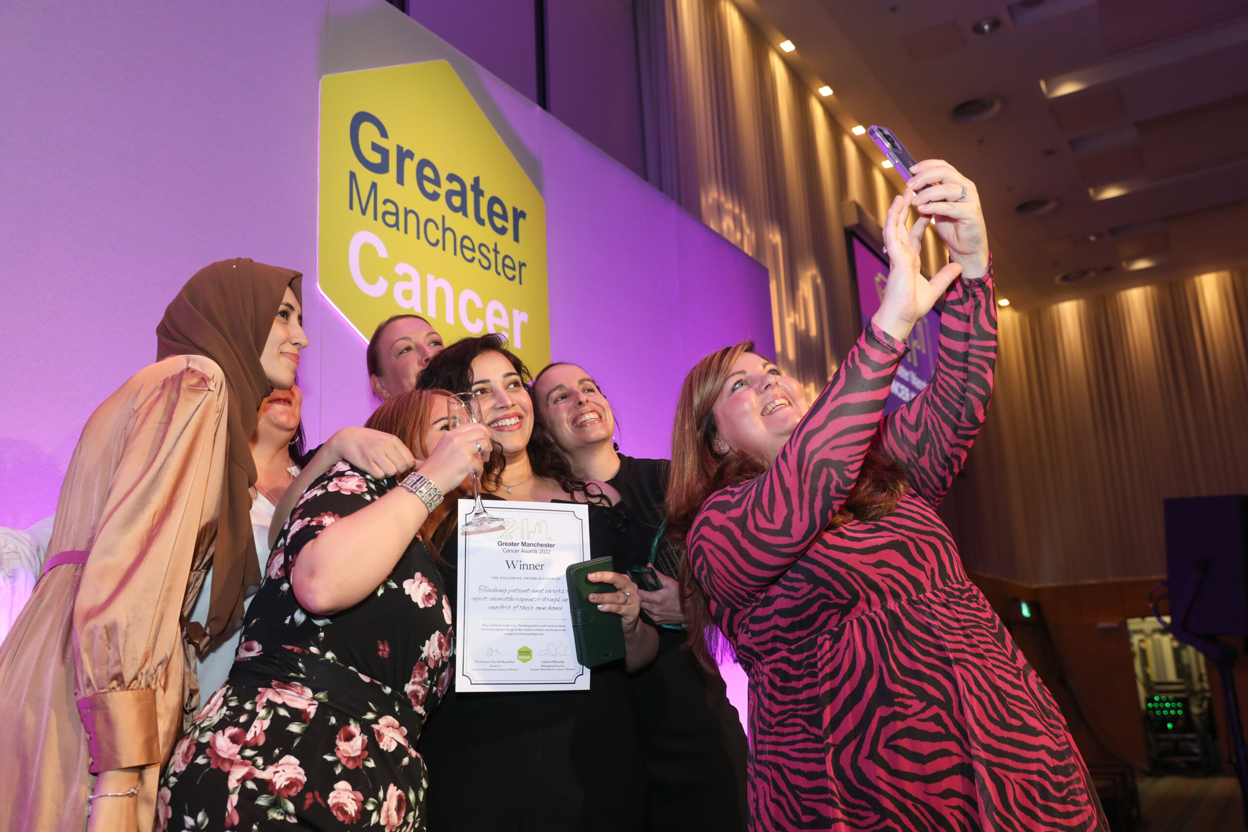 A woman in a red striped dress takes a selfie of colleagues on stage holding a 'winner' certificate