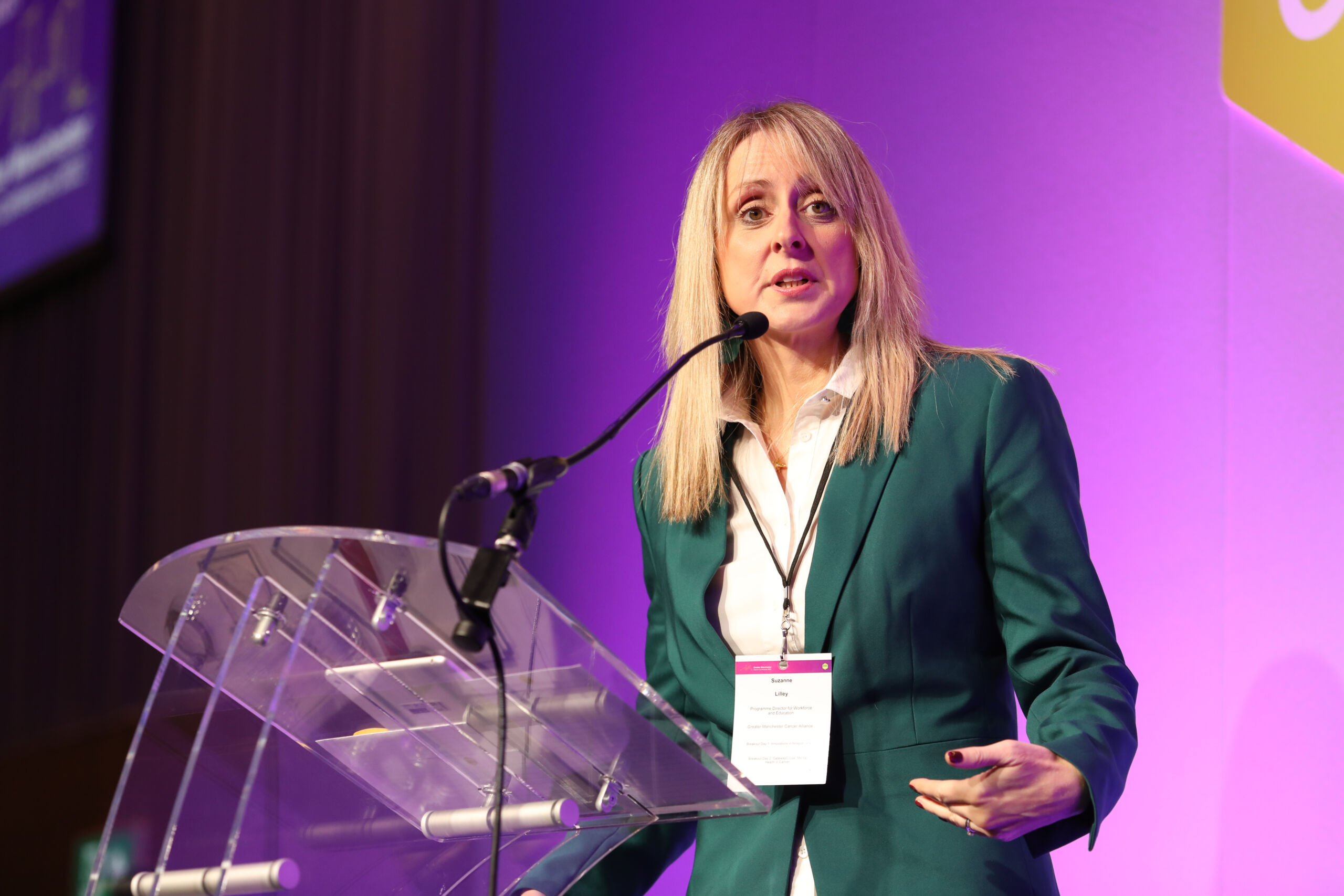 A white woman with blonde hair in a green suit talks at a lectern at Greater Manchester Cancer Conference