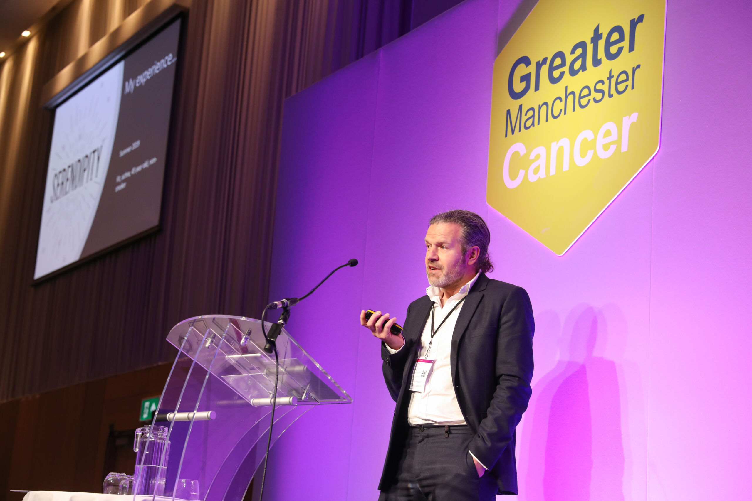 A white man in a dark suit talks at a lectern at Greater Manchester Cancer Conference 2022