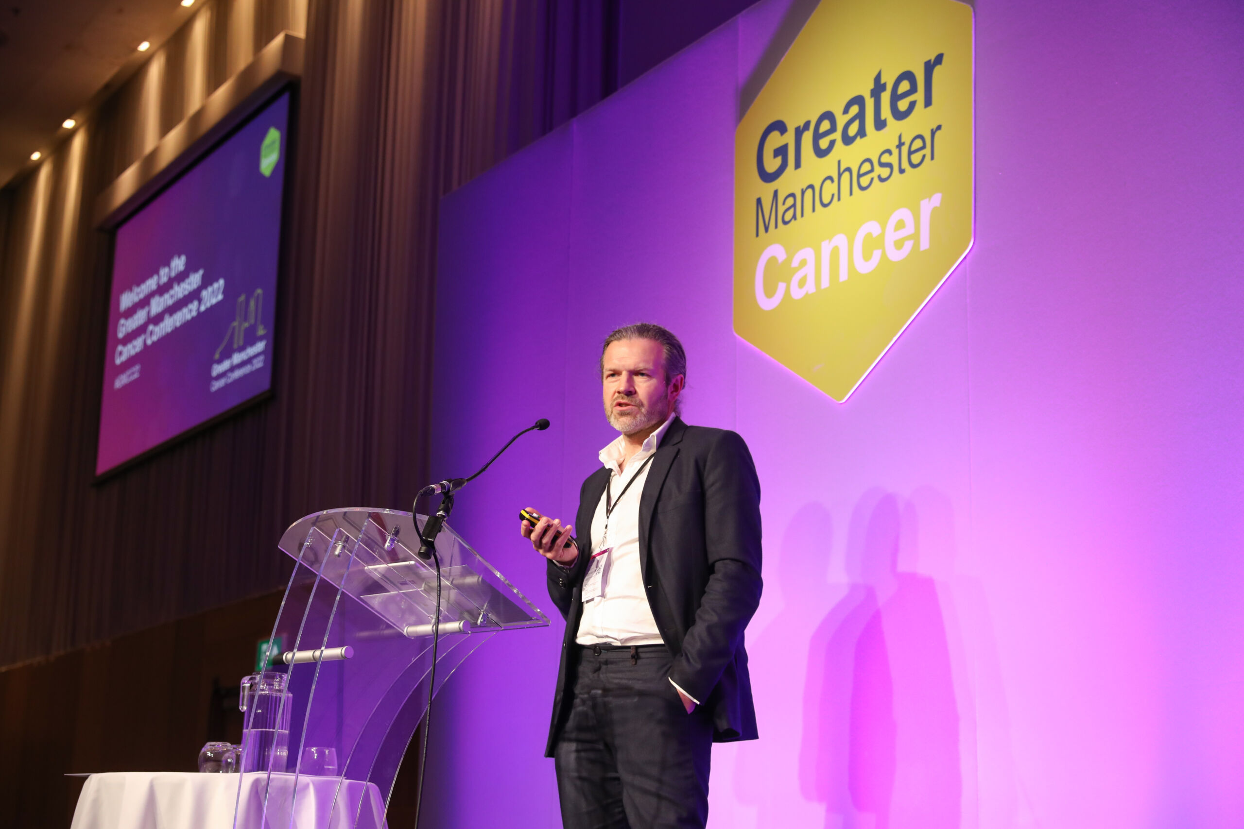 A white man in a dark suit talks at a lectern at Greater Manchester Cancer Conference 2022