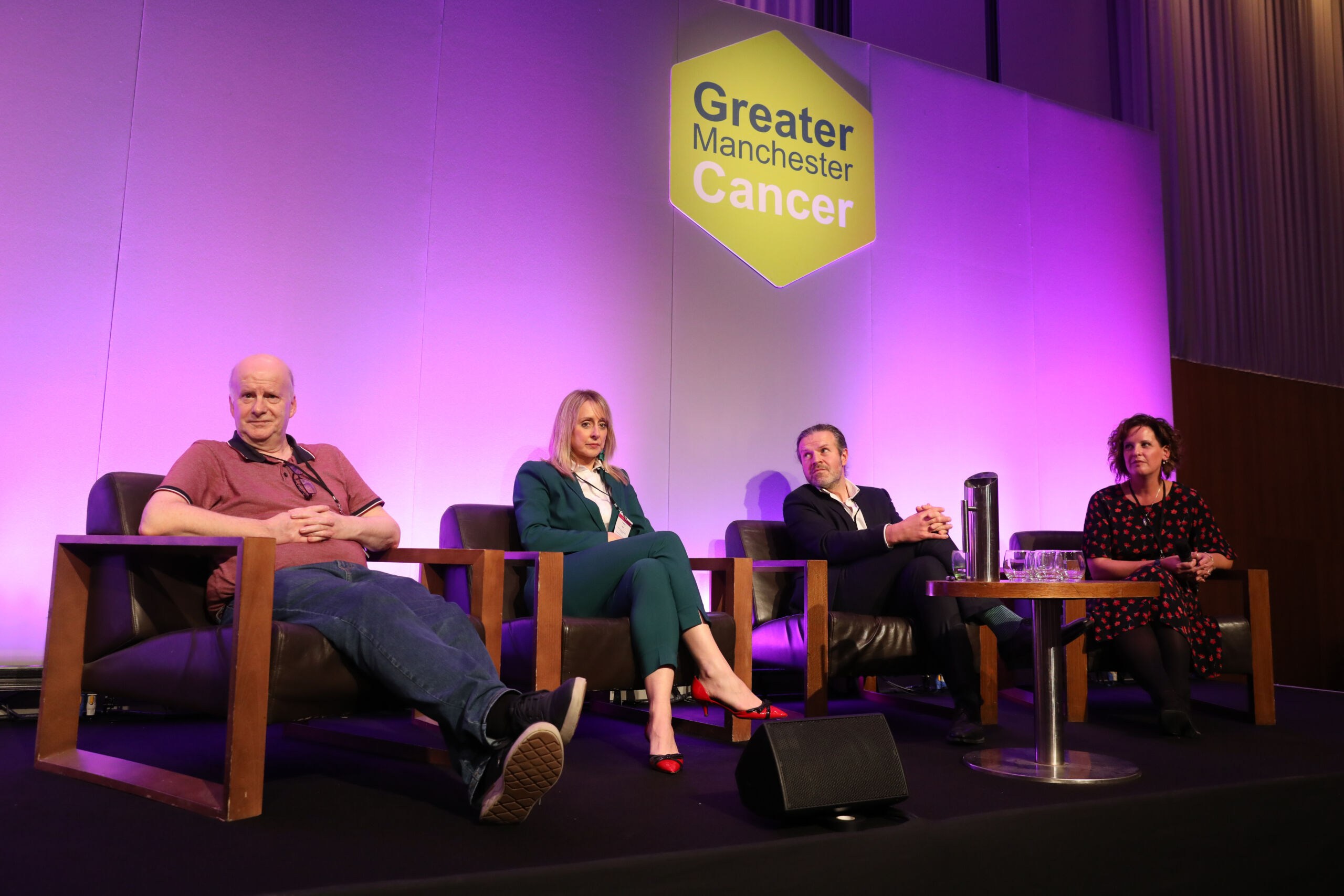 A panel of four speakers sit on low comfy chairs on stage with a green hexagon and the words Greater Manchester Cancer behind them.