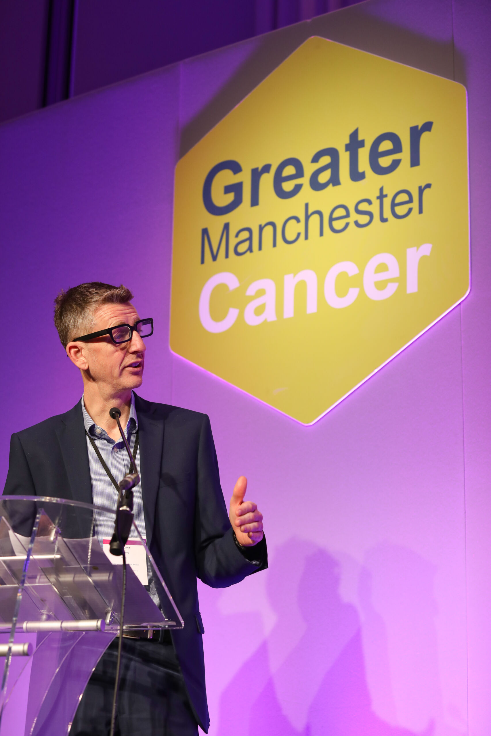 A white man in a dark suit stands on stage at a lectern in front of a green hexagon with the words Greater Manchester Cancer written on it