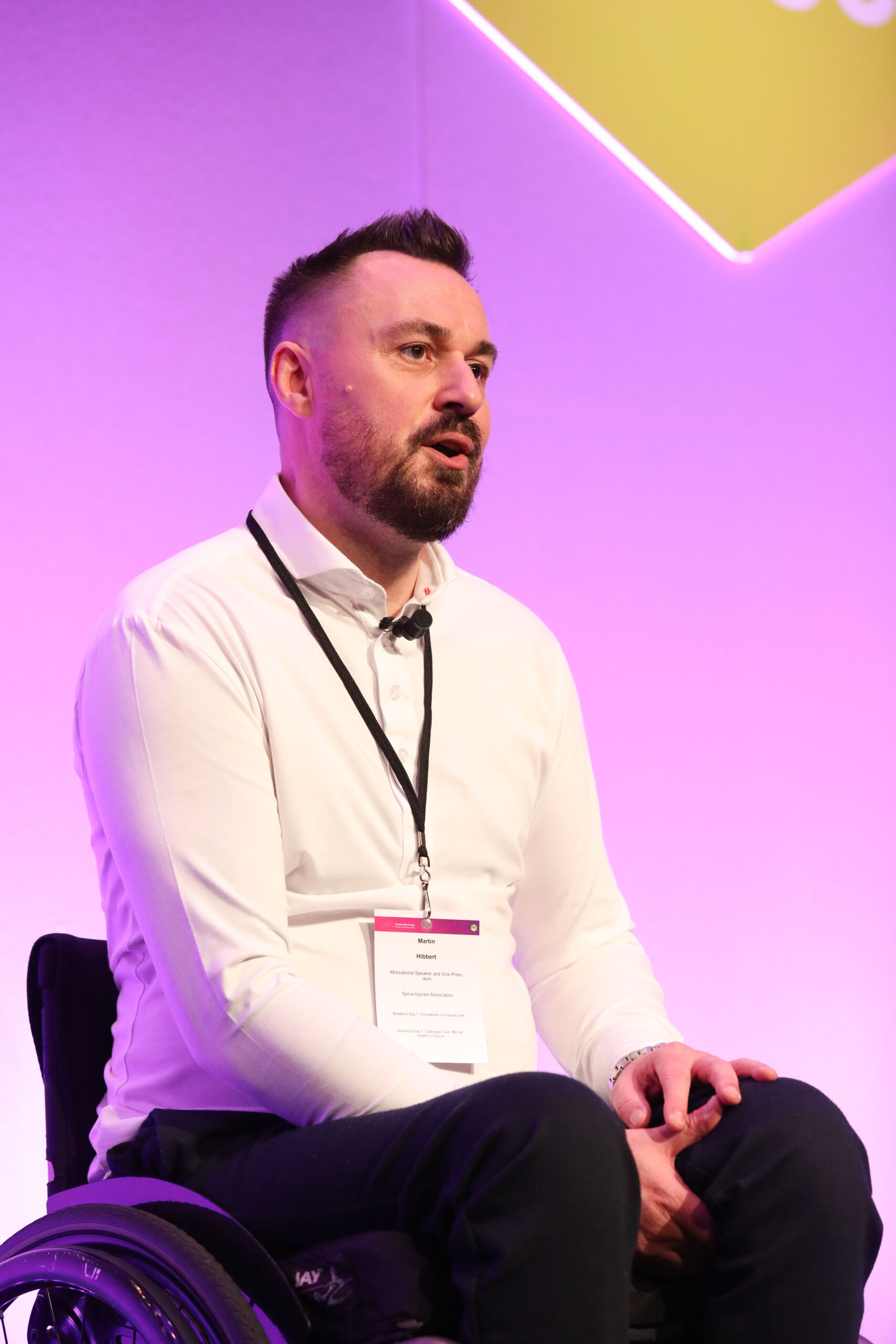 A close up head, shoulders and waist shot of a white man in a white shirt with dark hair and beards sitting on stage in a wheel chair at the Greater Manchester Cancer Conference