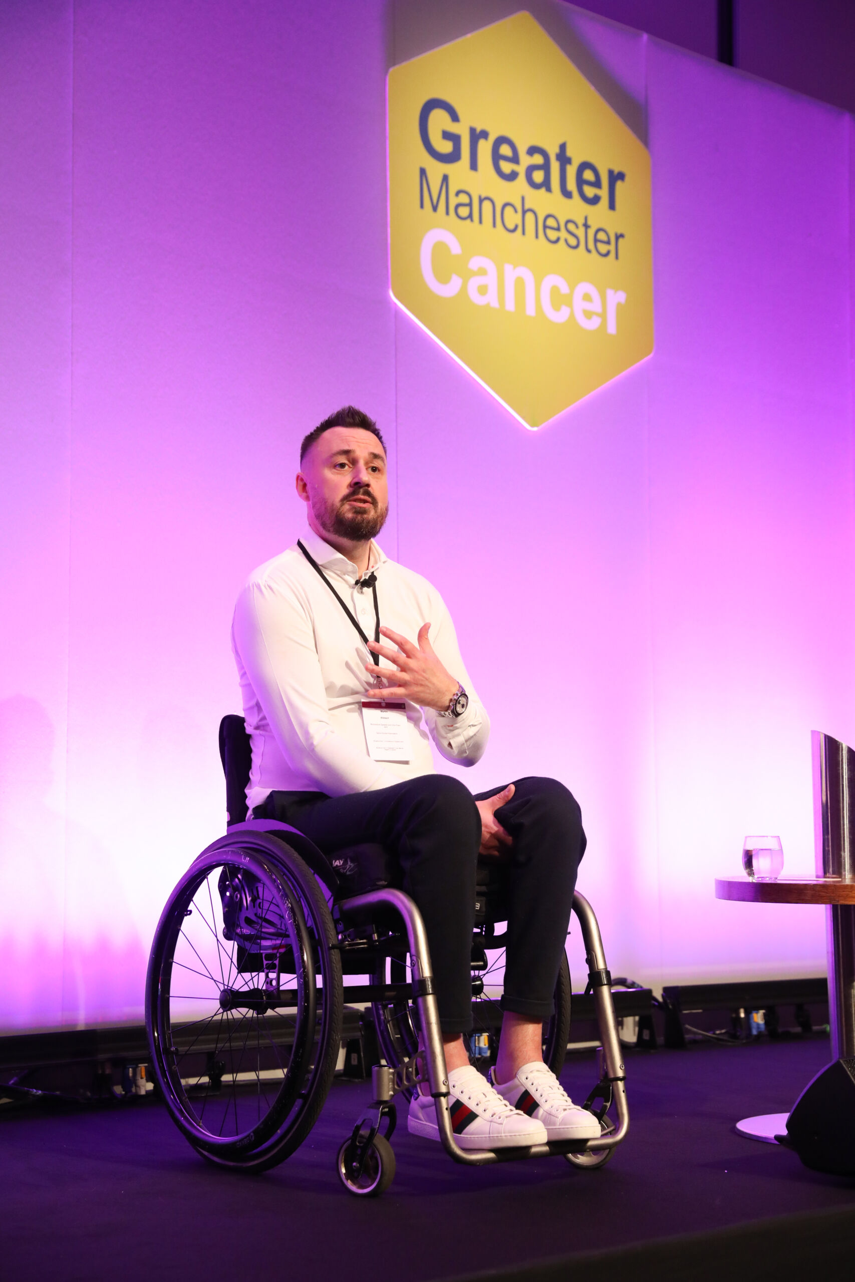 A white man in a white shirt with dark hair and beards sitting on stage in a wheel chair at the Greater Manchester Cancer Conference with the green Greater Manchester Cancer hexagon logo in the background