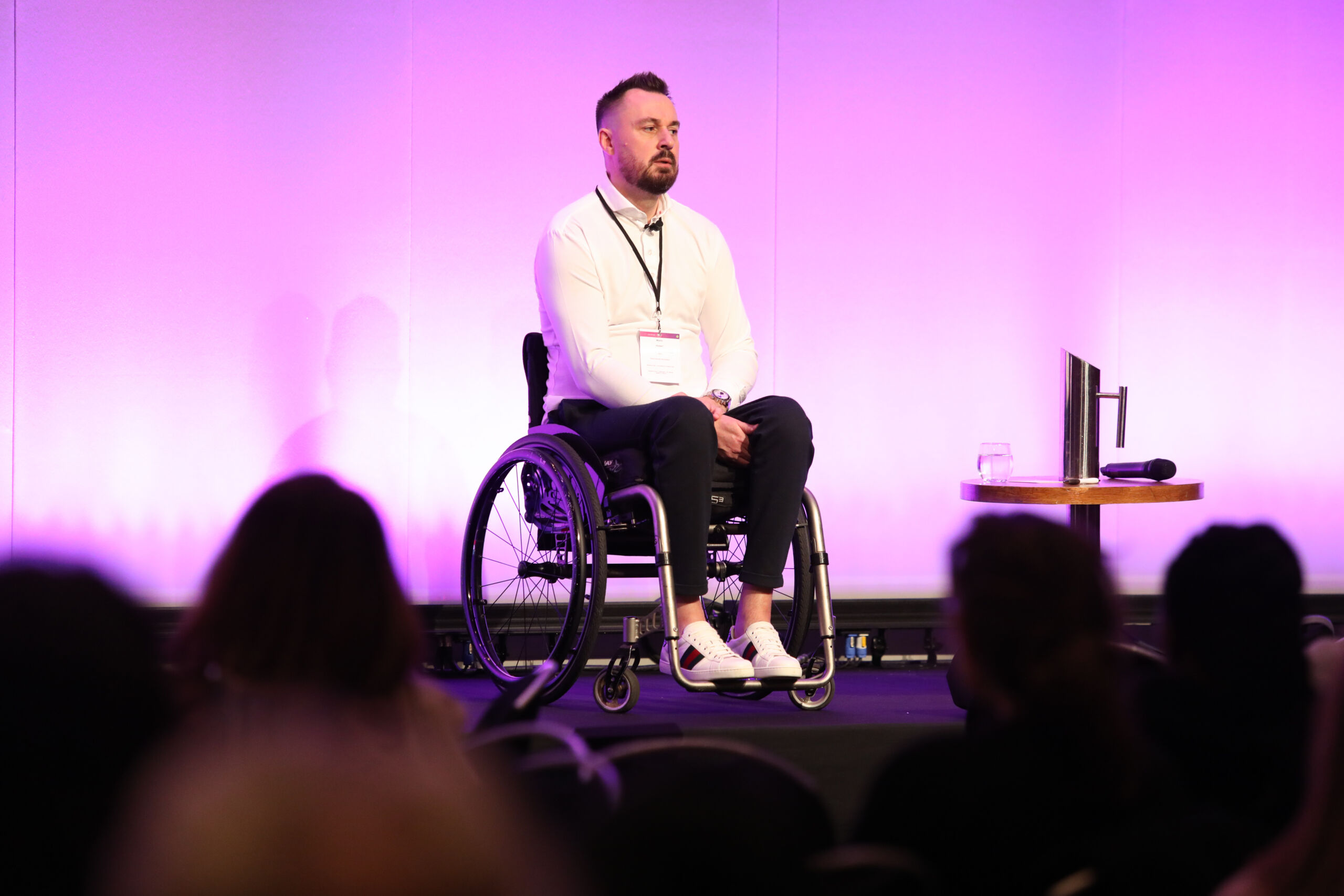 A white man in a white shirt with dark hair and beards sits in a wheel chair on stage at the Greater Manchester Cancer Conference. Heads from the audience can be seen in silhouette