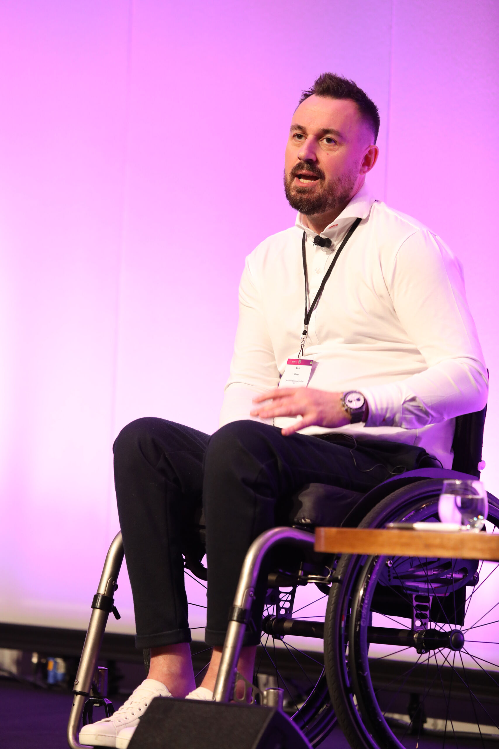 A white man in a white shirt with dark hair and beards sits in a wheel chair on stage at the Greater Manchester Cancer Conference