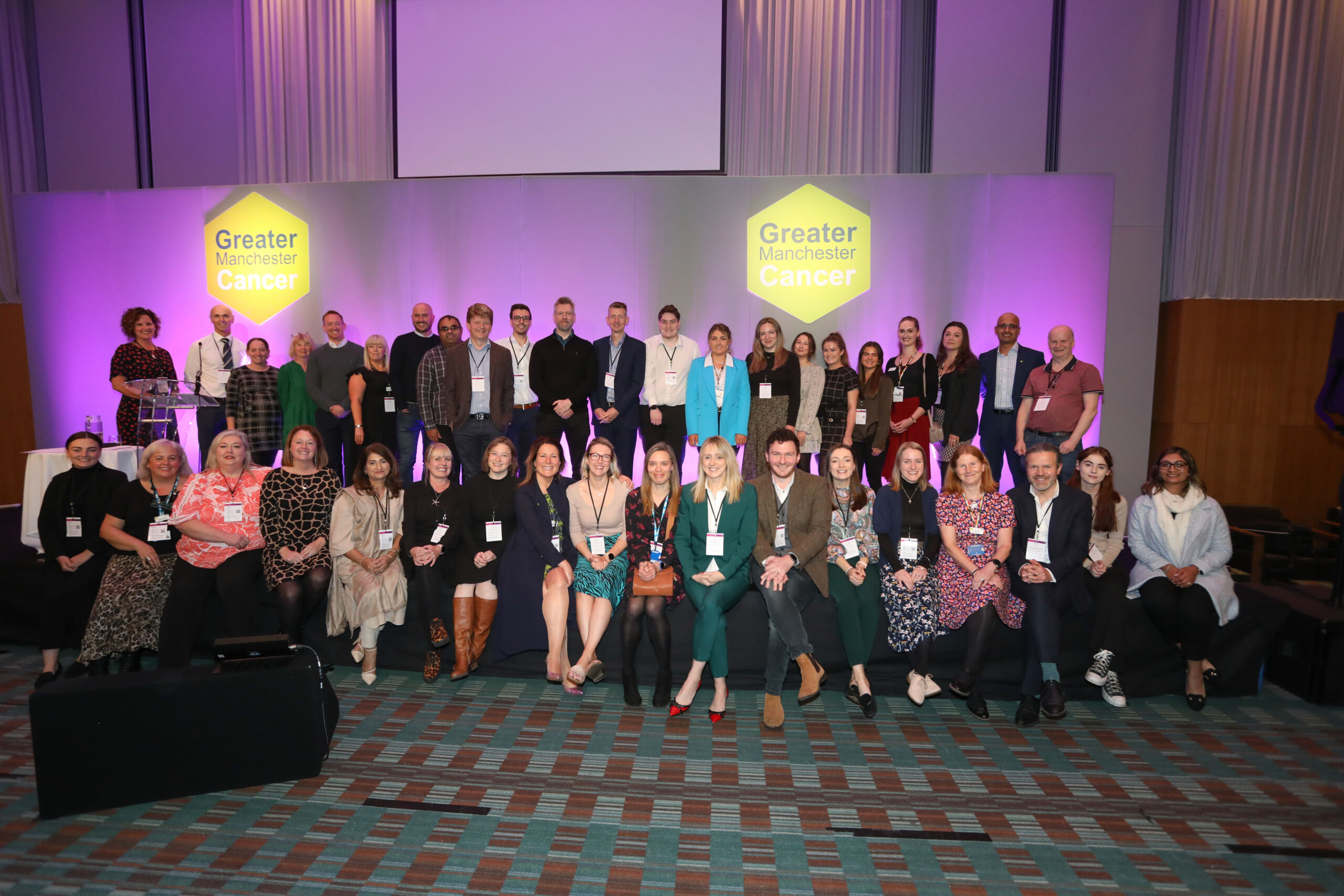 A group of around 40 men and women stand and sit on the stage at the Greater Manchester Cancer conference. There are two green hexagon logos with the words 'Greater Manchester Cancer' behind them.