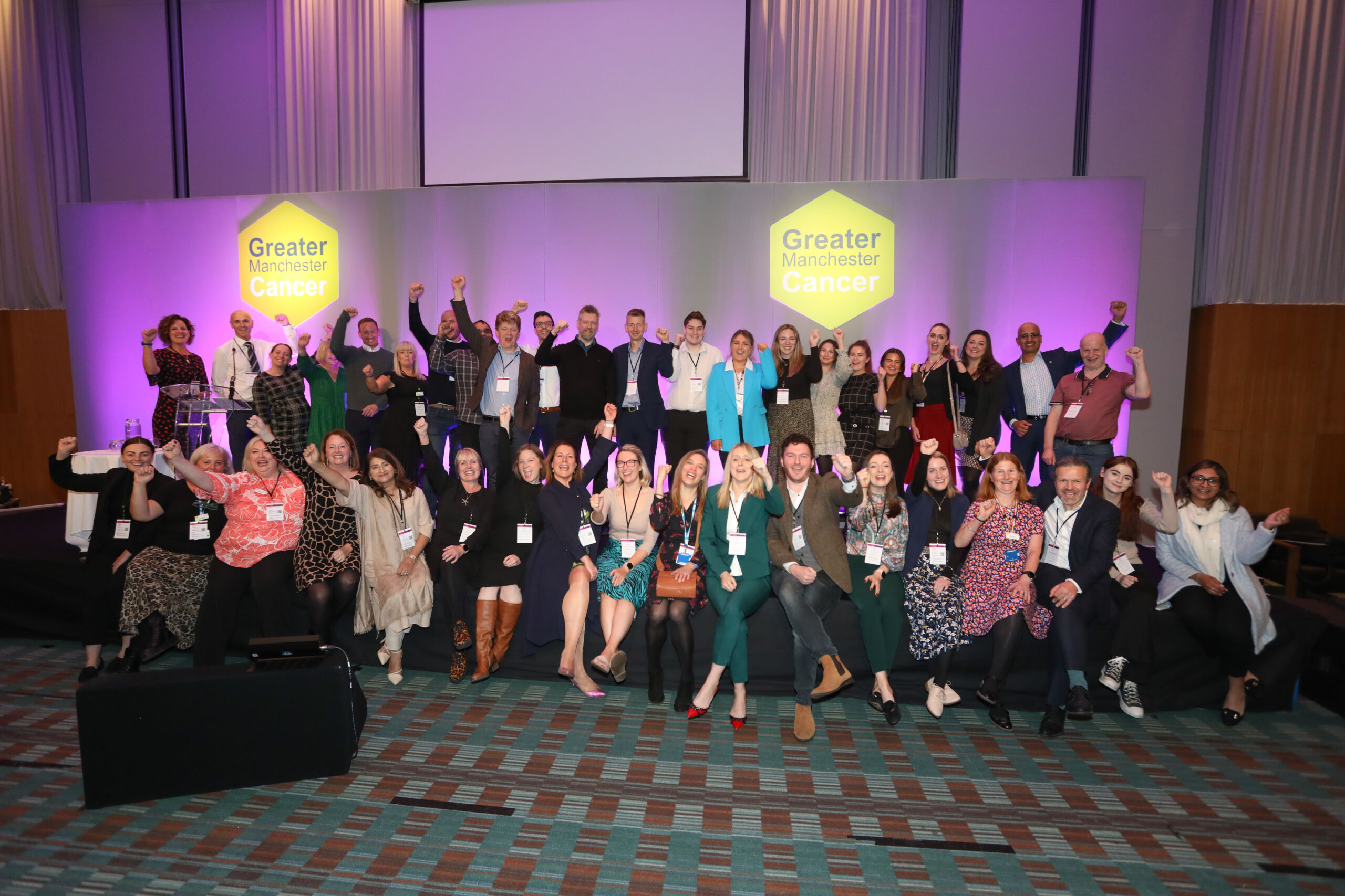 A group of around 40 people stand or sit on the stage at the Greater Manchester Cancer conference and raise their arms and cheer