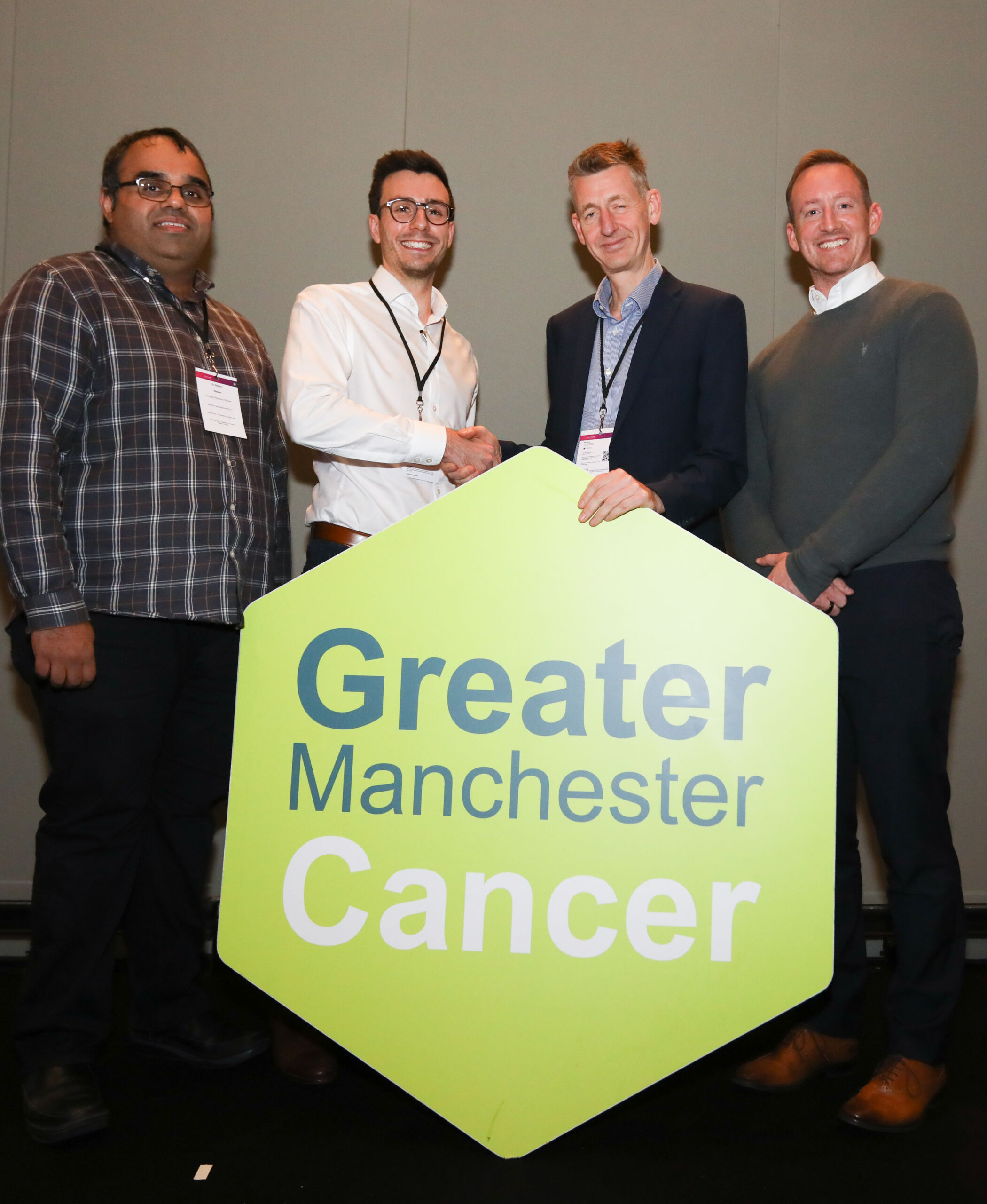Four men stand behind a giant green hexagon with the words Greater Manchester Cancer on it. The two in the centre are shaking hands.