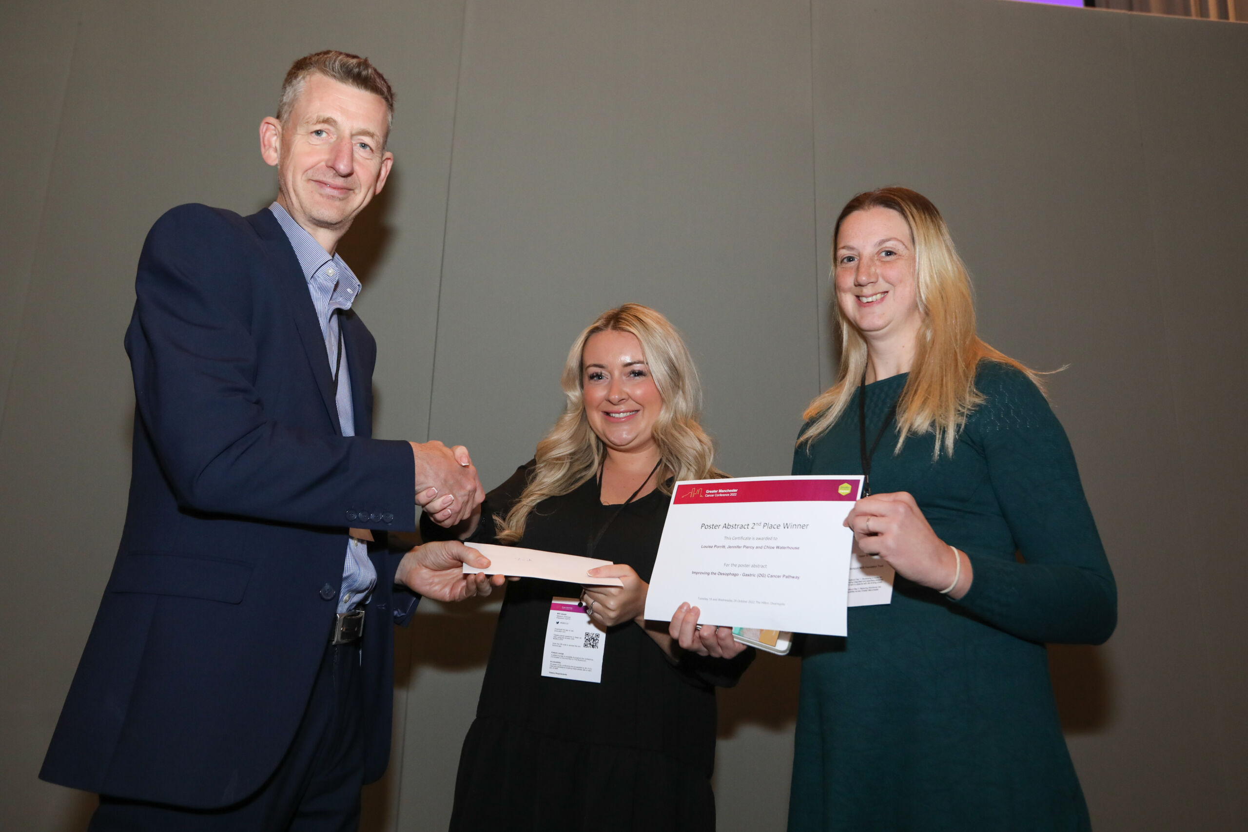 A man in a dark suit shakes hands with a woman with blonde hair while a second woman with blone hair holds a certifcatino saying Poster Awards Second Place Winner