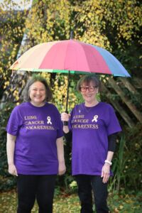 Two white women with grey hair wearing purple T-shirts and the words: Lung Cancer Awareness stand under a rainbow umbrella in a garden.