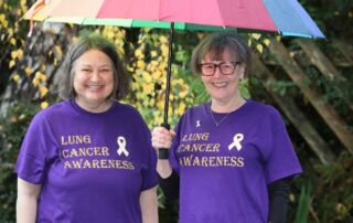 Two white women with grey hair wearing purple T-shirts and the words: Lung Cancer Awareness stand under a rainbow umbrella in a garden.