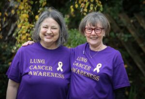 Two white women with grey hair one shoulder length one in a bob stand next to each other with their arms around each other in a green garden. They wear purple T-shirts with the words Lung Cancer Awareness written on them.