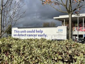 The long white NHS Galleri unit with the words 'This unit could help us detect cancer early' is parked in Salford near a green hedge