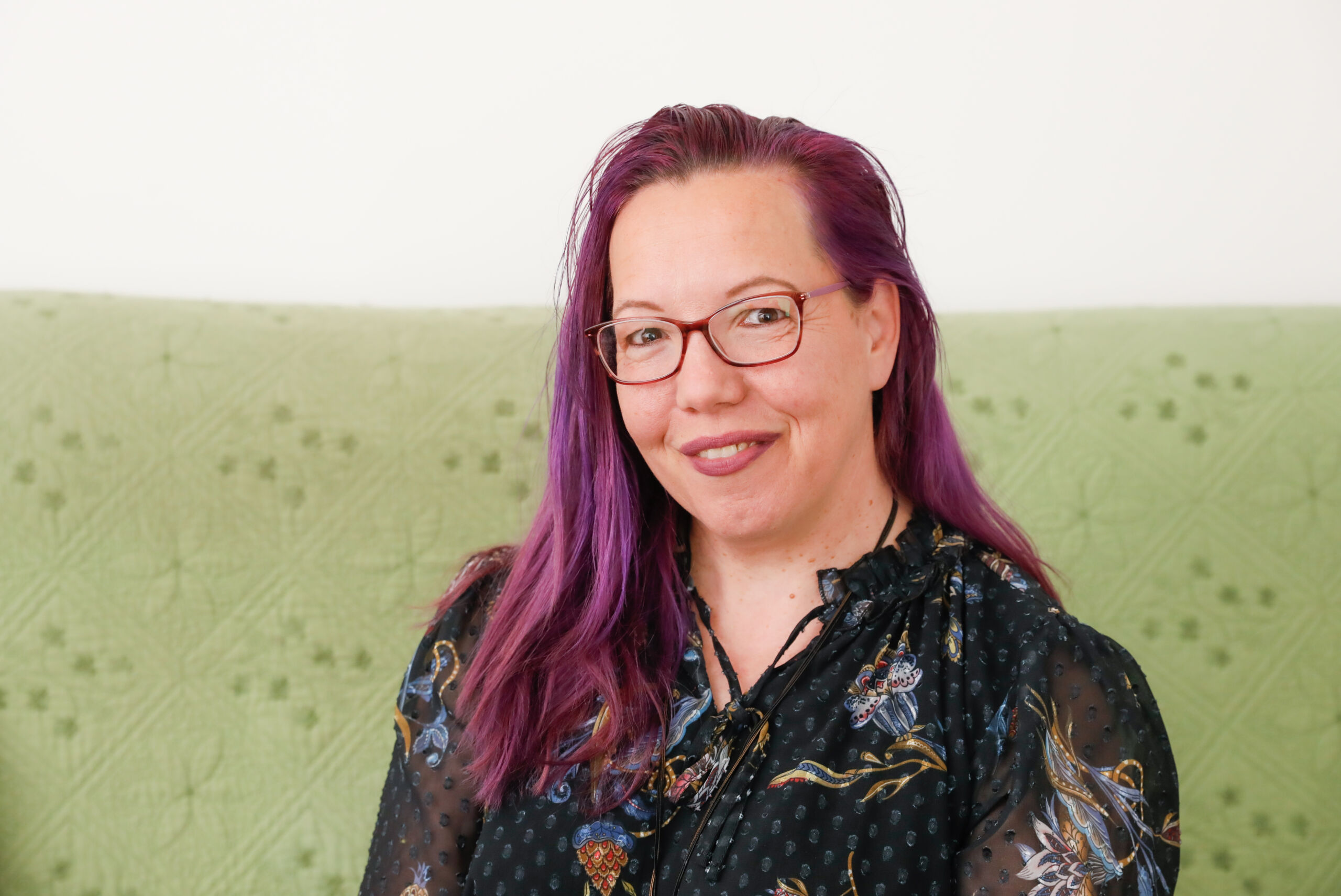 Mihaela Atkinson a white woman with long purple hair sits on a green sofa wearing pink glasses