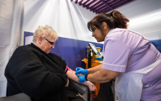 A patient - a white woman with blonde hair and glasses has his blood sample taken by a female Asian nurse with black hair tied in a pony tail