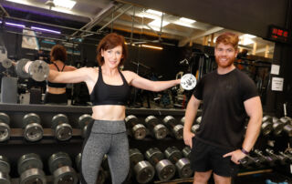 Tracey a white woman with shortish brown hair holds weights outstretch in a gym as her personal trainer Thomas a white man with red hair stands by her side