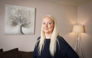 Laura Hope a white woman with long blonde hair stands in front of a tree picture on a wall in her living room in Oldham