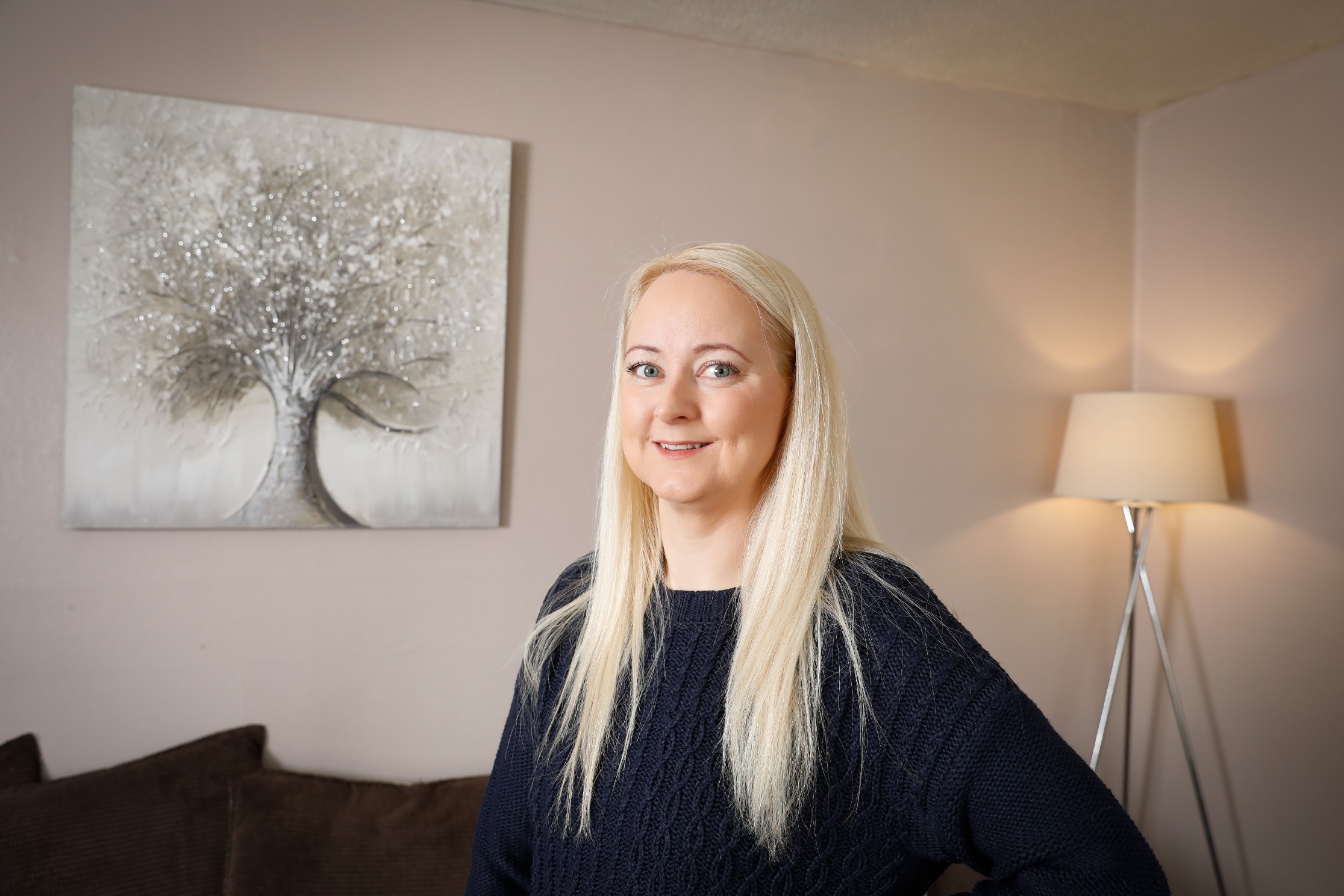 Laura Hope a white woman with long blonde hair stands in front of a tree picture on a wall in her living room in Oldham