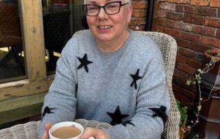 Gill Broom a white woman with short white hair wearing glasses and a grey jumper with black stars on it sitting at a table with a mug of coffee