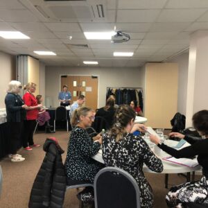 Alison Foxley, Communications and Engagement Manager and Head judge Miss Susannah Penney, Associate Director at Greater Manchester Cancer Alliance (standing) address a room full of people sat at round tables