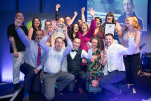 Members of the Targeted Lung Health Check team who won the Patients' Choice Award with their arms in the air celebrating on stage at the Greater Manchester Cancer Awards