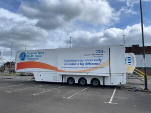 A large white truck housing the Targeted Lung Health Check truck parked in a car park in Greater Manchester. Words on the side of the truck are the words: "Targeted Lung Health Check Programme" and "Catching lung cancer early can make a big difference. For more information please come inside and talk to our team."