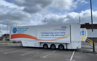 A large white truck housing the Targeted Lung Health Check truck parked in a car park in Greater Manchester. Words on the side of the truck are the words: "Targeted Lung Health Check Programme" and "Catching lung cancer early can make a big difference. For more information please come inside and talk to our team."