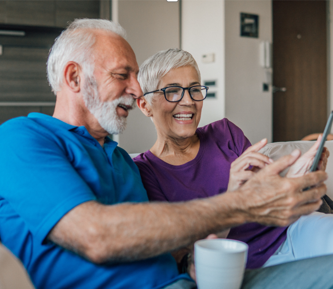 a man and woman sit on a sofa, using a phone or ipad. They are both smiling.