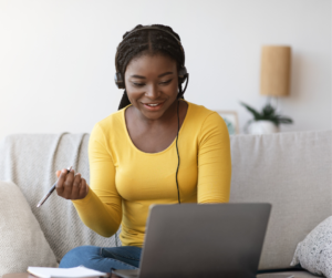 A person sits on a couch with a laptop on their lap, wearing headphones and holding a pen, smiling while looking at the screen. There are papers and a notebook beside them, suggesting they are engaged in a study or work session.