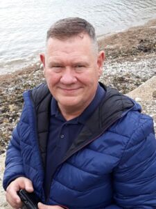 Person smiling at the camera, wearing a blue jacket, with a beach in the background.
