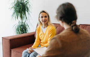 A white woman in a yellow jumper sitting on a comfy sofa talking to a counsellor