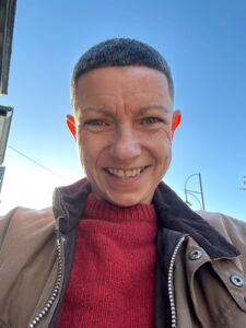 A selfie image of Sharon a lady in her mid-40s with short dark grey hair wearing a red top and brown jacket with a bright blue sky behind her.