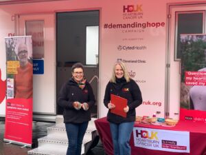Two women stand in front of a white roadshow unit with a table with leaflets nearby and the Heartburn Cancer UK's logo on the front.