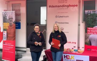 Two women stand in front of a white roadshow unit with a table with leaflets nearby and the Heartburn Cancer UK's logo on the front.