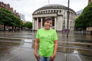 A Black woman in her 40s stands in front of central reference library in Manchester city centre wearing a green T-shirt with a hand and an eye on the front to encourage people to look and feel their breasts