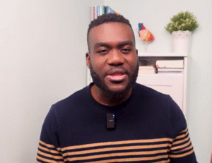 Dr Derek, a young black man in a GP surgery with a set of shelves behind him.
