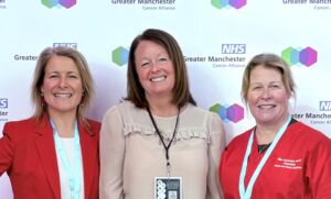 GM Cancer leadership2 Three individuals wearing lanyards stand smiling in front of a backdrop with the NHS and Greater Manchester logos. Two are wearing red tops and one is in a beige blouse.