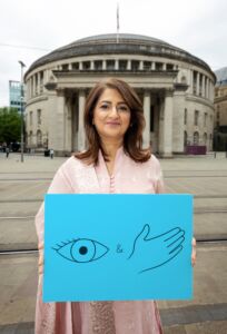 Nabila Farooq who features in the Look and Feel Monthly campaign to raise awareness about checking your breasts Nabila Farooq standing with a blue sign featuring an eye and hand in front of Manchester Library.