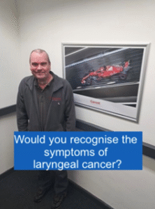 Person in a black uniform standing in front of a framed photo of a red Formula 1 car with a caption about recognizing the symptoms of laryngeal cancer.
