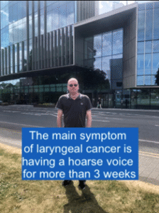 Person standing in front of a modern building with a sign displaying a health message about laryngeal cancer symptoms.