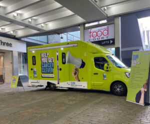 A green van with the words This Van Can Help You Get Cancer Clever written on the side parked in Market Street Manchester in front of shops