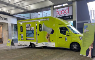 A green van with the words This Van Can Help You Get Cancer Clever written on the side parked in Market Street Manchester in front of shops