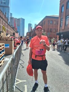 Tom - an older white man with grey hair wears and orange T-shirt and and cap and running kit and holds a medal at the finish line of the Manchester 10K